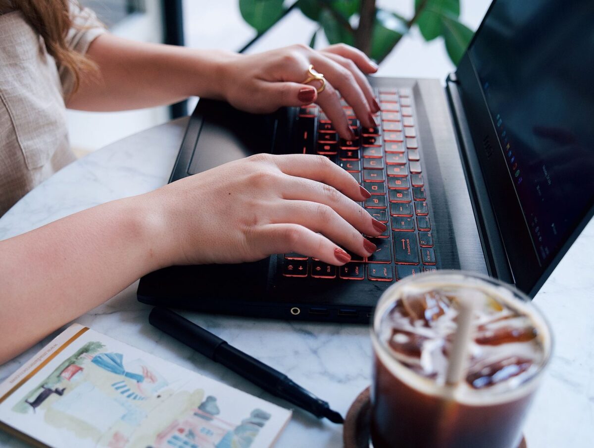 A writer working on a laptop at a café table with a notebook and iced coffee