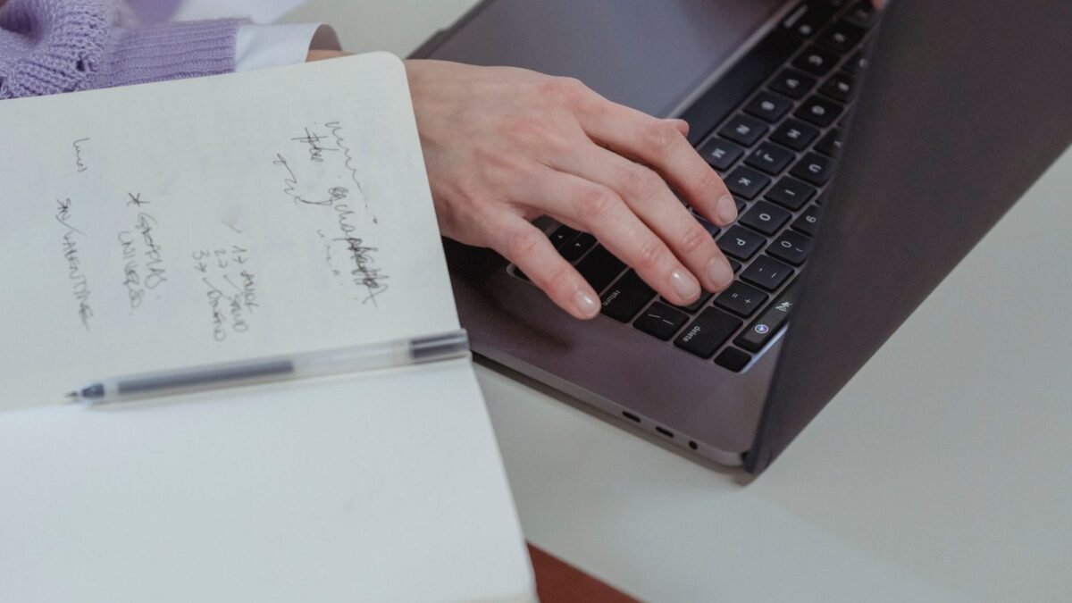 An editor typing on a laptop with an open handwritten notebook beside her